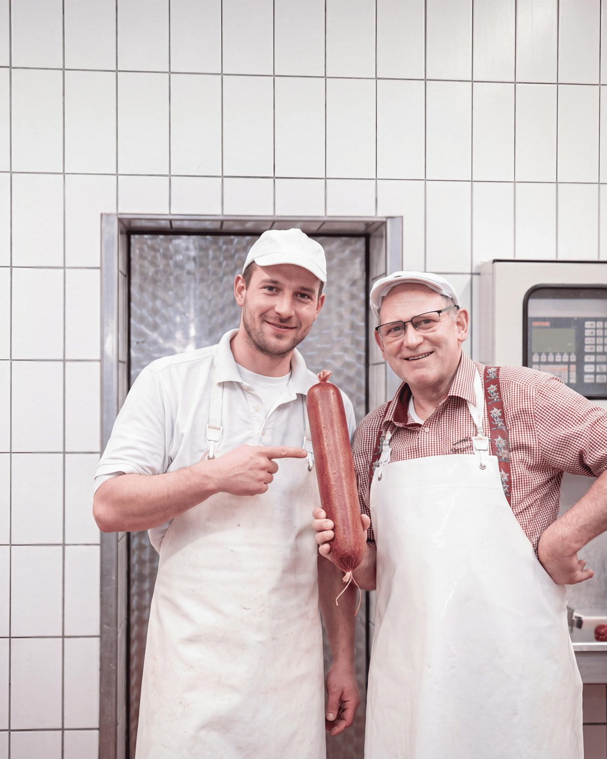 Zwei Männer in weißen Schürzen und Kappen stehen in einer Metzgerei, einer hält und beide zeigen auf eine große Wurst.